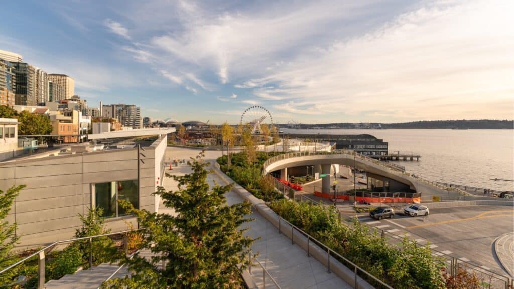 The Seattle Waterfront boasts modern buildings, lush green spaces, and a transformative ferris wheel near the shoreline, all under a partly cloudy sky. The Overlook Walk offers stunning views of this vibrant urban scene.