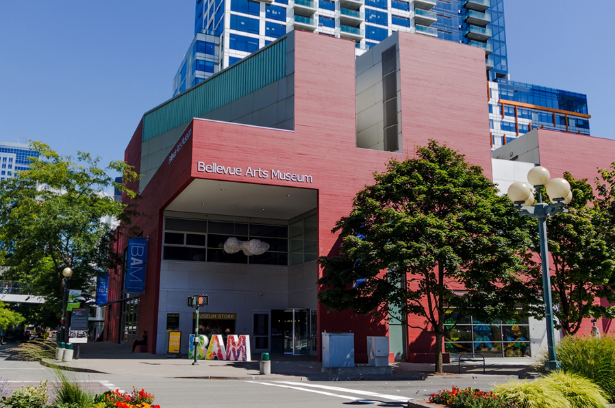 Outdoor view of the Bellevue Arts Museum's modern exterior with red and glass facade, located alongside trees and a street with crosswalk and streetlights.